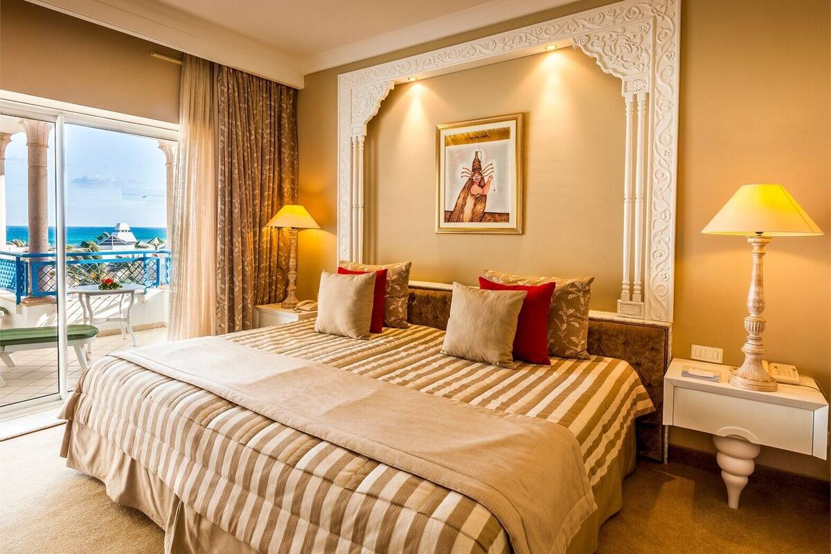 Een hotelkamer met een groot bed, twee nachtkastjes, uitzicht op zee en een balkon met een tafel en stoelen.