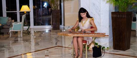 A woman seated on a chair playing a stringed instrument in a well-lit lobby with marble flooring, large windows, and potted plants.