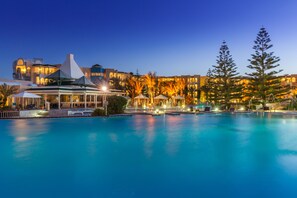 Un resort con una amplia piscina, varias edificaciones con iluminación cálida y un árbol destacado al frente.
