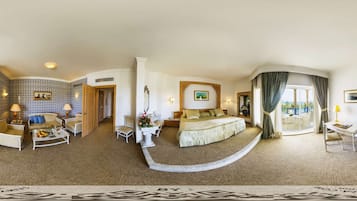 Amplia habitación de hotel con una cama grande, zona de estar con sofá y sillones, mesa de comedor y vistas al exterior.
