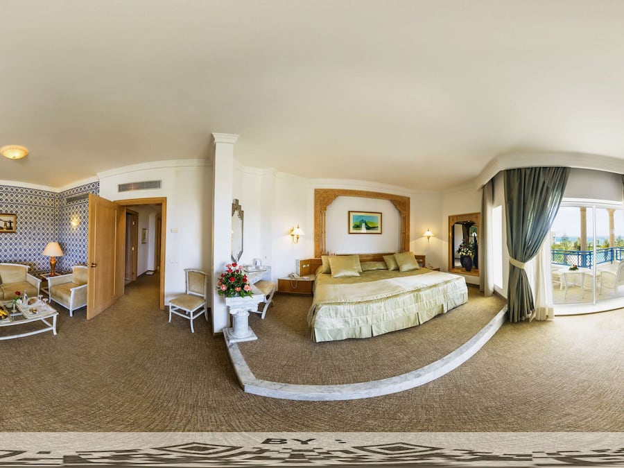 Amplia habitación de hotel con una cama grande, zona de estar con sofá y sillones, mesa de comedor y vistas al exterior.