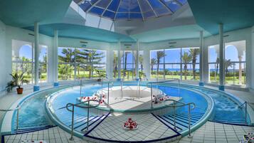 Una amplia piscina interior con un jacuzzi central, rodeada por una terraza con baldosas y grandes ventanales que ofrecen vistas a palmeras y al océano.