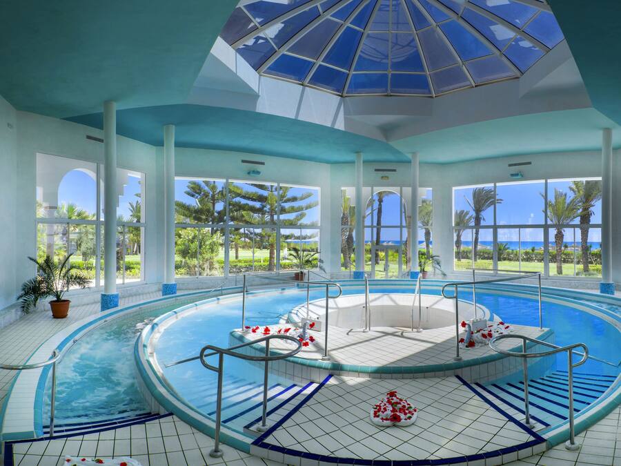 Una amplia piscina interior con un jacuzzi central, rodeada por una terraza con baldosas y grandes ventanales que ofrecen vistas a palmeras y al océano.