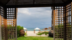 Blick auf einen Strand durch eine Holzgitterstruktur, mit Sandstrand und zwei Gazebos mit Reetdach.
