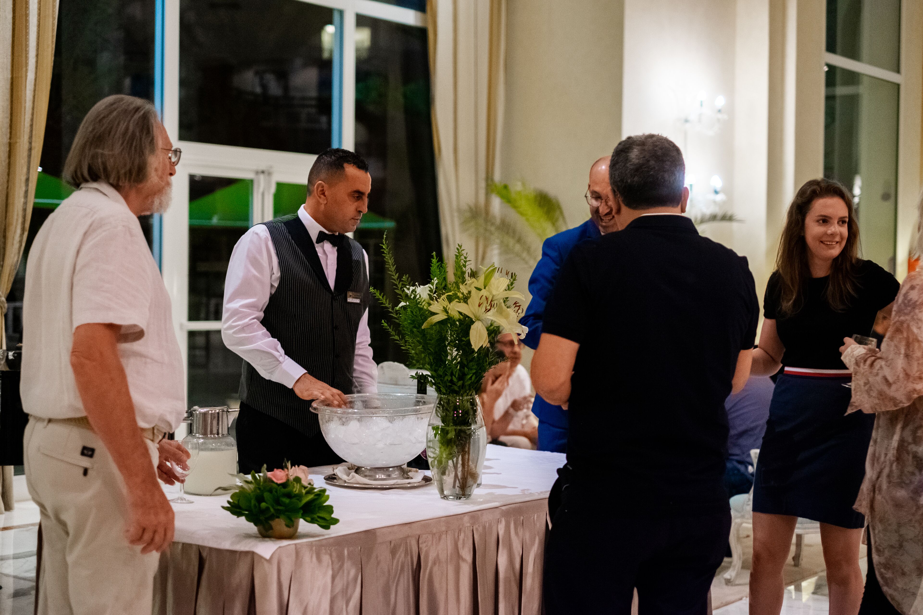 Un grupo de personas en una zona de recepción con una mesa de buffet, un jarrón con flores y un tazón grande de hielo.