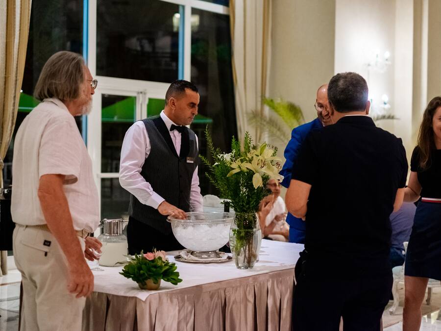 Un grupo de personas en una zona de recepción con una mesa de buffet, un jarrón con flores y un tazón grande de hielo.