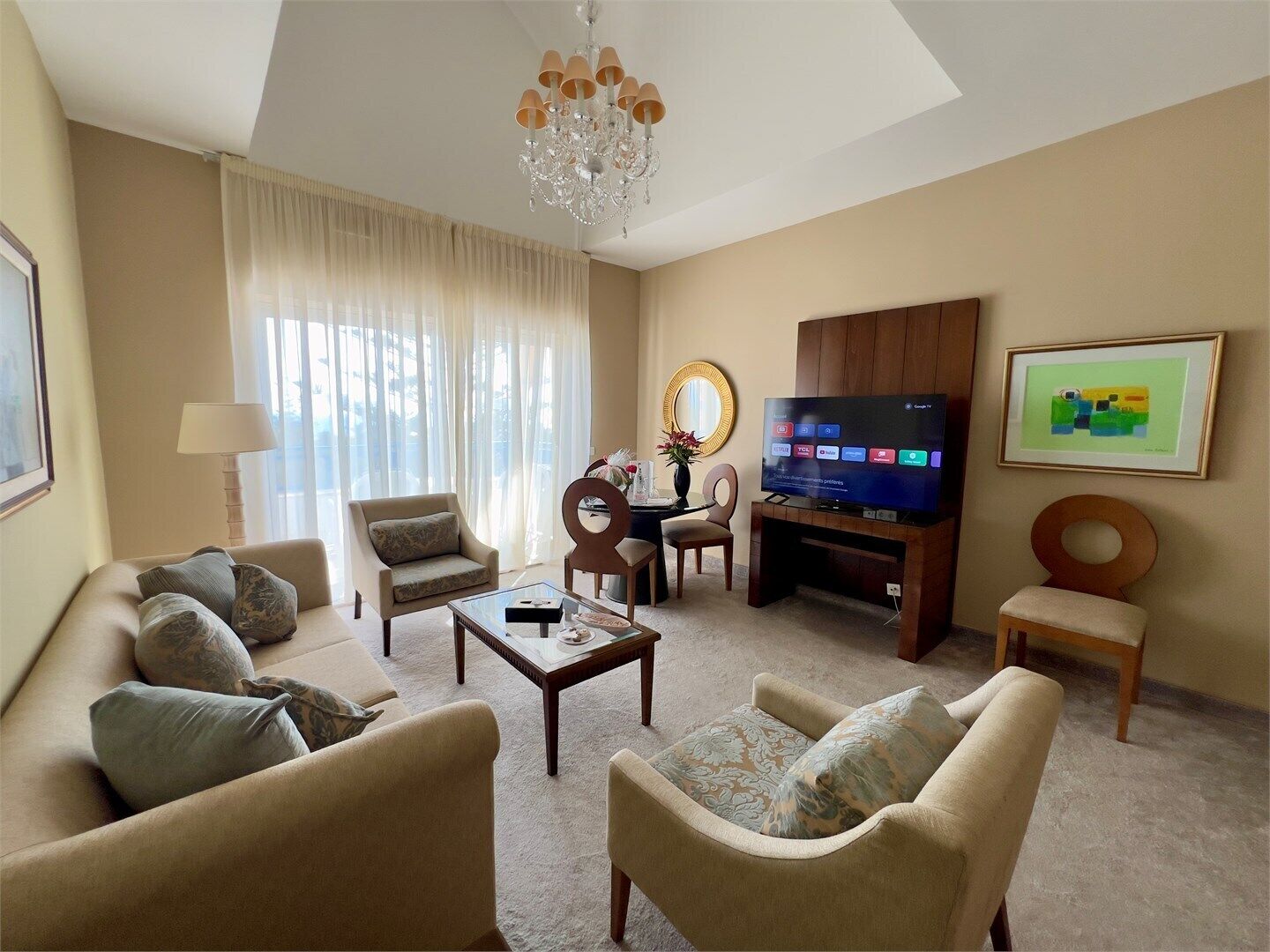 Ein Wohnzimmer mit einer Couch, Sesseln, einem Couchtisch, einem Fernseher, einer Kristallleuchtre und einem gerahmten Bild an der Wand.