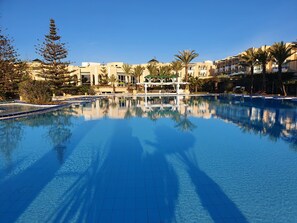 Una gran piscina cristalina con solárium de baldosas, rodeada de palmeras y edificios con balcones.