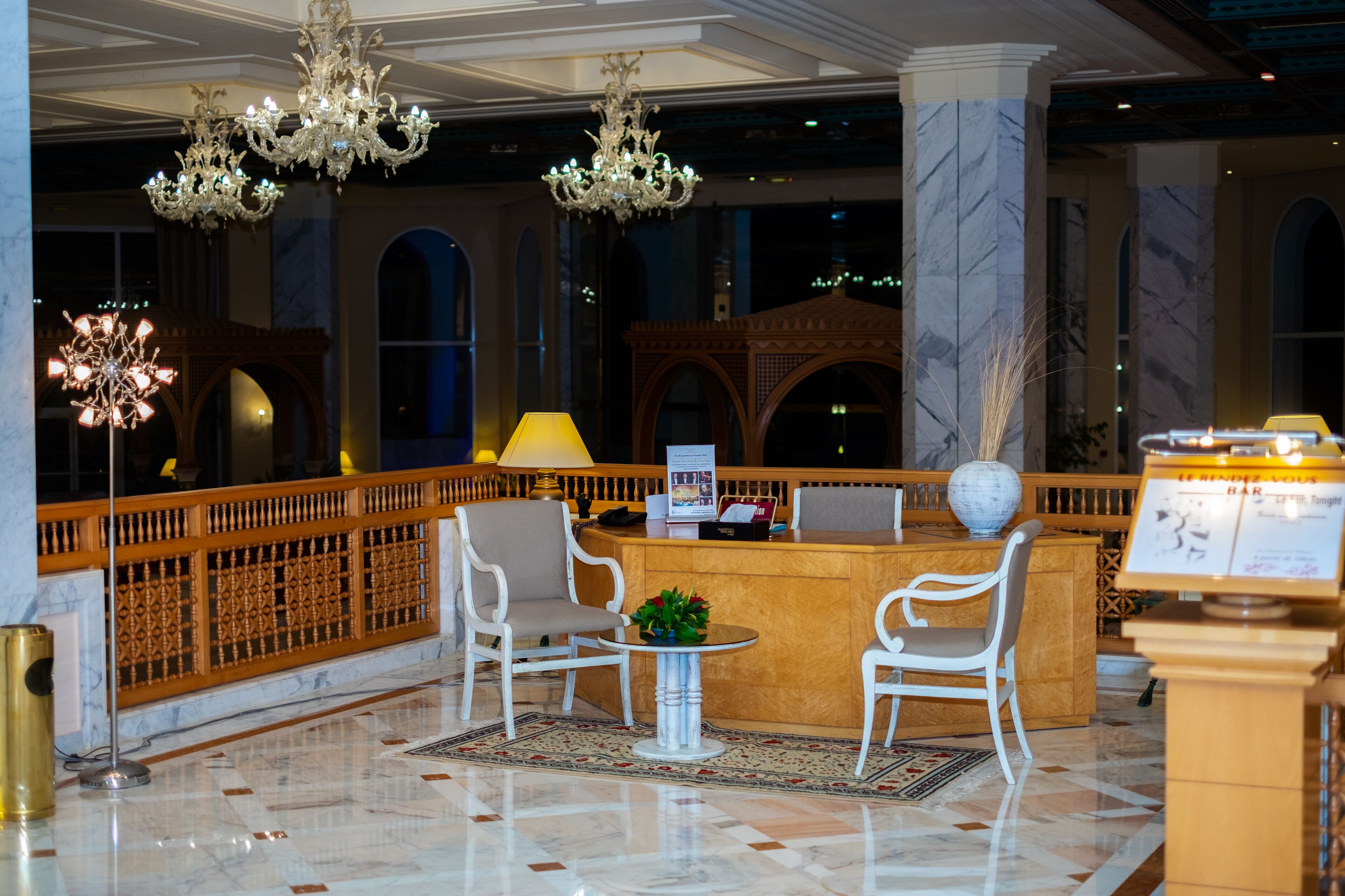 Un vestíbulo con mostrador de recepción, sillas blancas, candelabros y suelo de mármol.