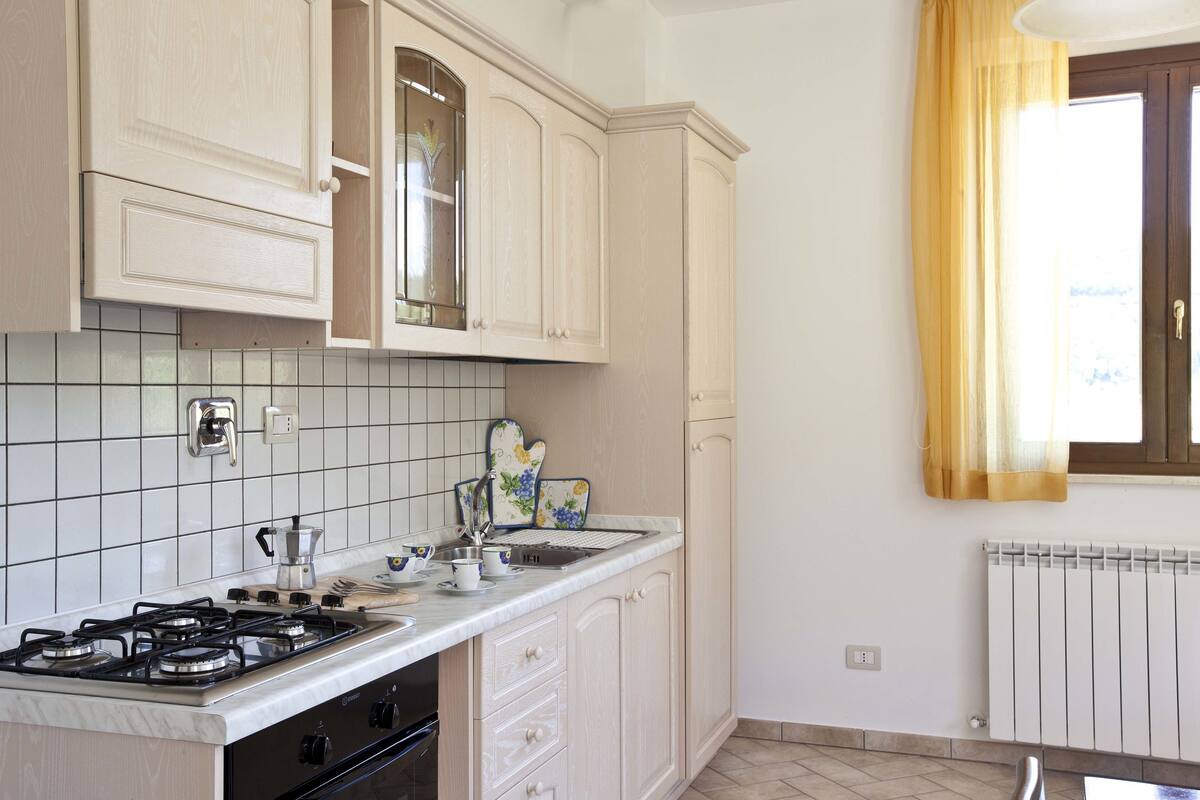 A kitchen with white cabinets, a gas stove, a sink, and a window with yellow curtains.