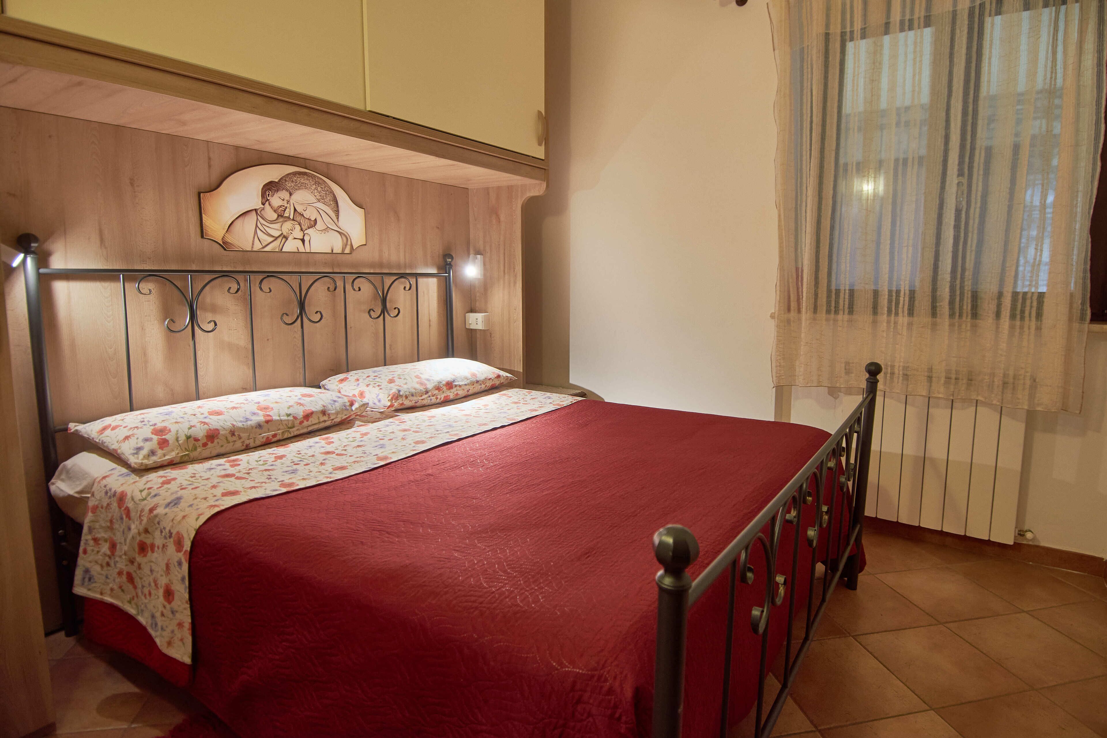 Un lit avec une couvre-lit rouge et des coussins à motifs floraux, une tête de lit en bois ornée de sculptures, et une fenêtre avec des rideaux légers.