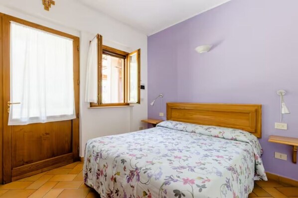 Un dormitorio con cama de madera, ventana con cortinas, mesita de noche y lámpara de pared.