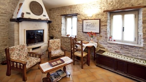 A cozy living room with stone walls, a fireplace, and a television. There are two floral armchairs and a wooden coffee table with books. - Il Castello (Assisi)