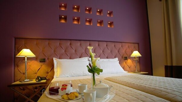A hotel room with a tufted headboard, two beds, bedside lamps, a tray with breakfast items, and a wall with recessed lighting.