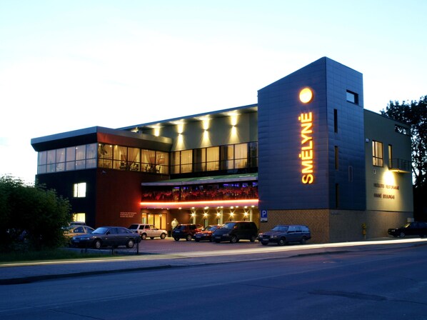 A modern building with a prominent 'SMELYNE' sign, featuring a large glass facade and a parking area in front.