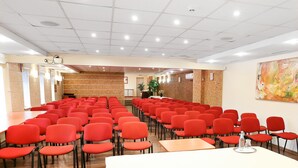 Konferenssihuone, jossa on riveissä punaisia tuoleja ja pitkä pöytä. Lattia on puuta ja taustaseinässä on korkkipintaa.