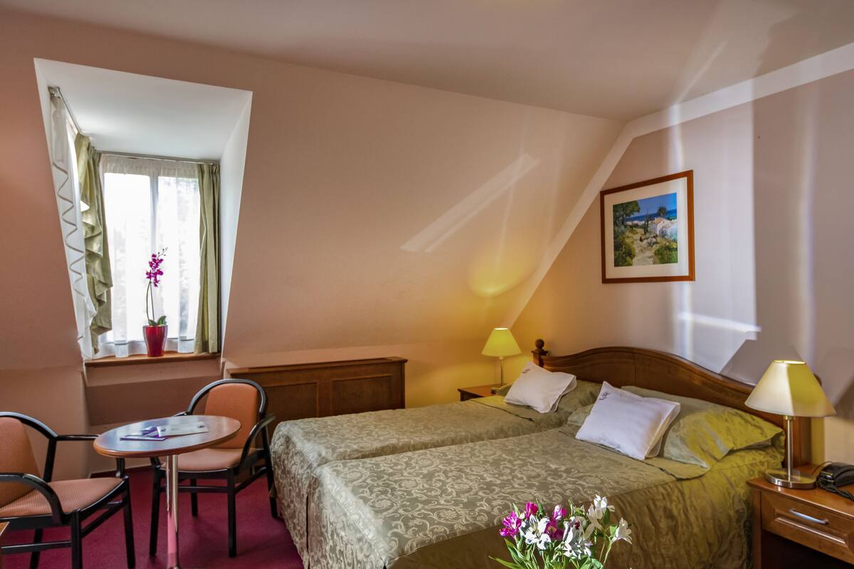 A hotel room with two beds, a small table with chairs, a window with curtains, a bedside table with a lamp, and a framed picture on the wall.