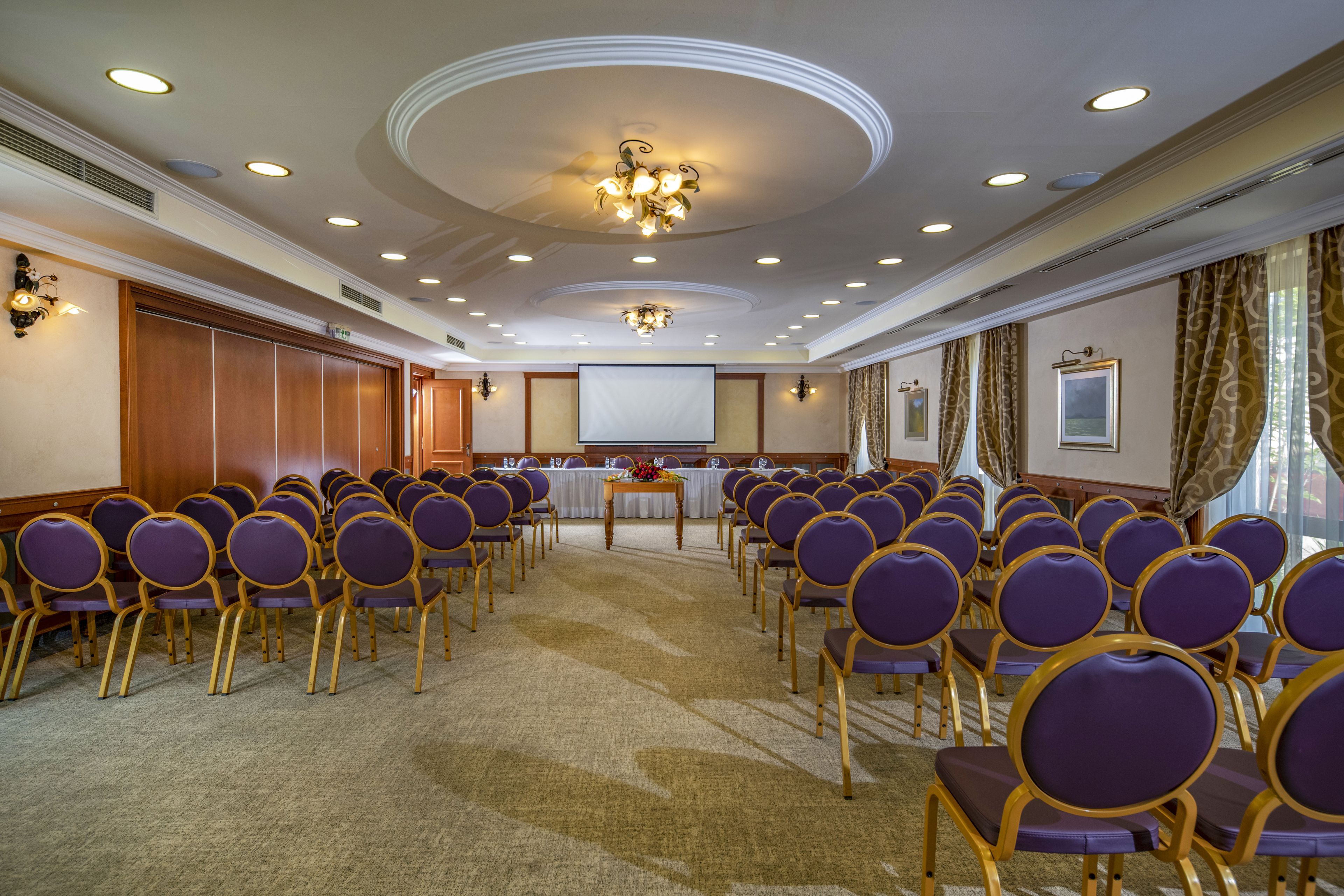 A conference room with rows of purple chairs, a projector screen, and a wooden table with a floral arrangement.