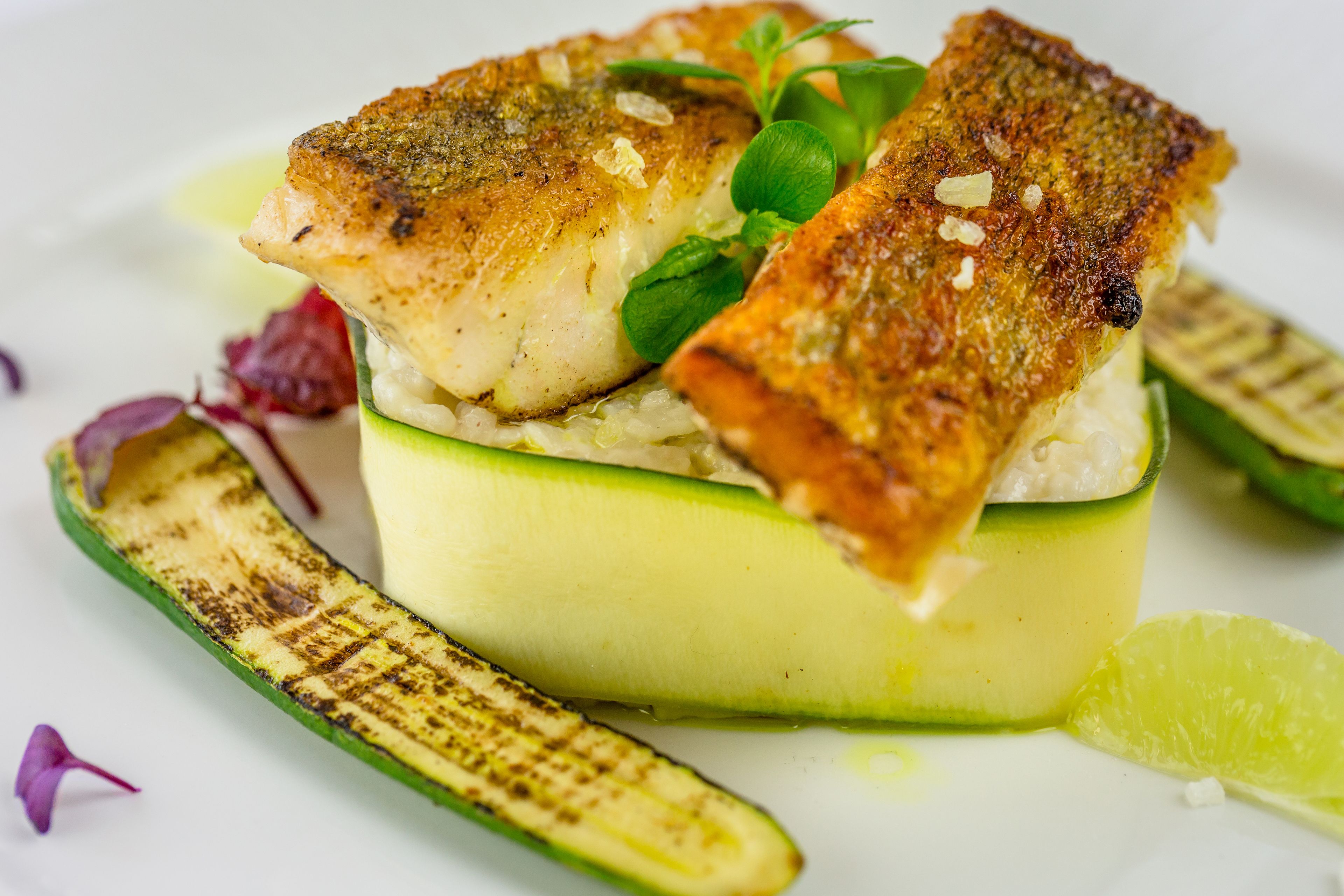 Un plato con pescado a la parrilla sobre una base de verduras verdes, acompañado de calabacín a la parrilla y una rodaja de lima.