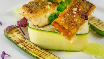Un plato con pescado a la parrilla sobre una base de verduras verdes, acompañado de calabacín a la parrilla y una rodaja de lima.