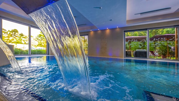 Una piscina interior con caída de agua, ventanas amplias y vista a la vegetación exterior.