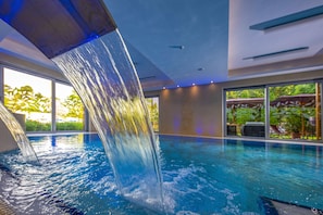 An indoor swimming pool with a waterfall feature, large windows, and a view of greenery outside. - Betekints Wellness és Konferencia Hotel (Veszprem)