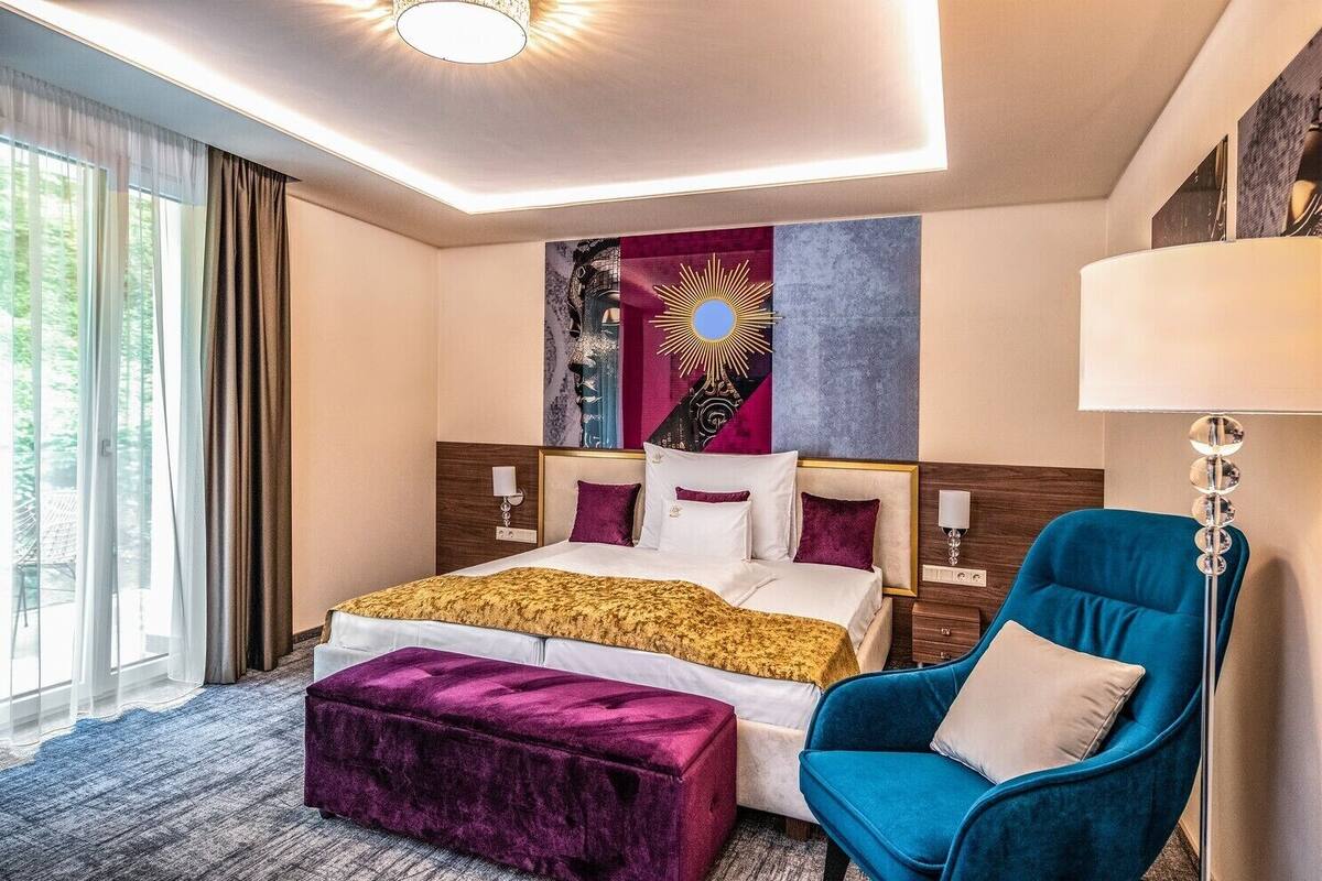 A modern hotel room with a large bed, a blue armchair, and a view of greenery outside.