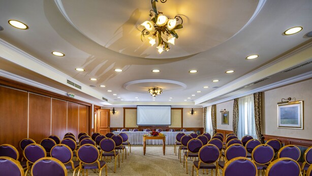 A conference room with rows of purple chairs, a projector screen, and a table with floral arrangements. - Betekints Wellness és Konferencia Hotel (Veszprem)