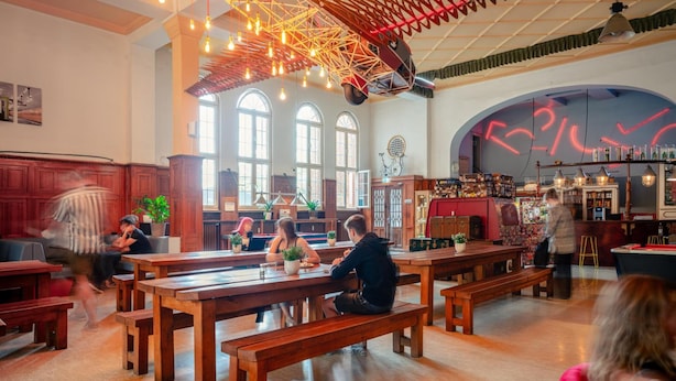 Un espace intérieur spacieux, doté de bancs et de tables en bois, de grandes fenêtres et d’un plafond orné de luminaires suspendus.
