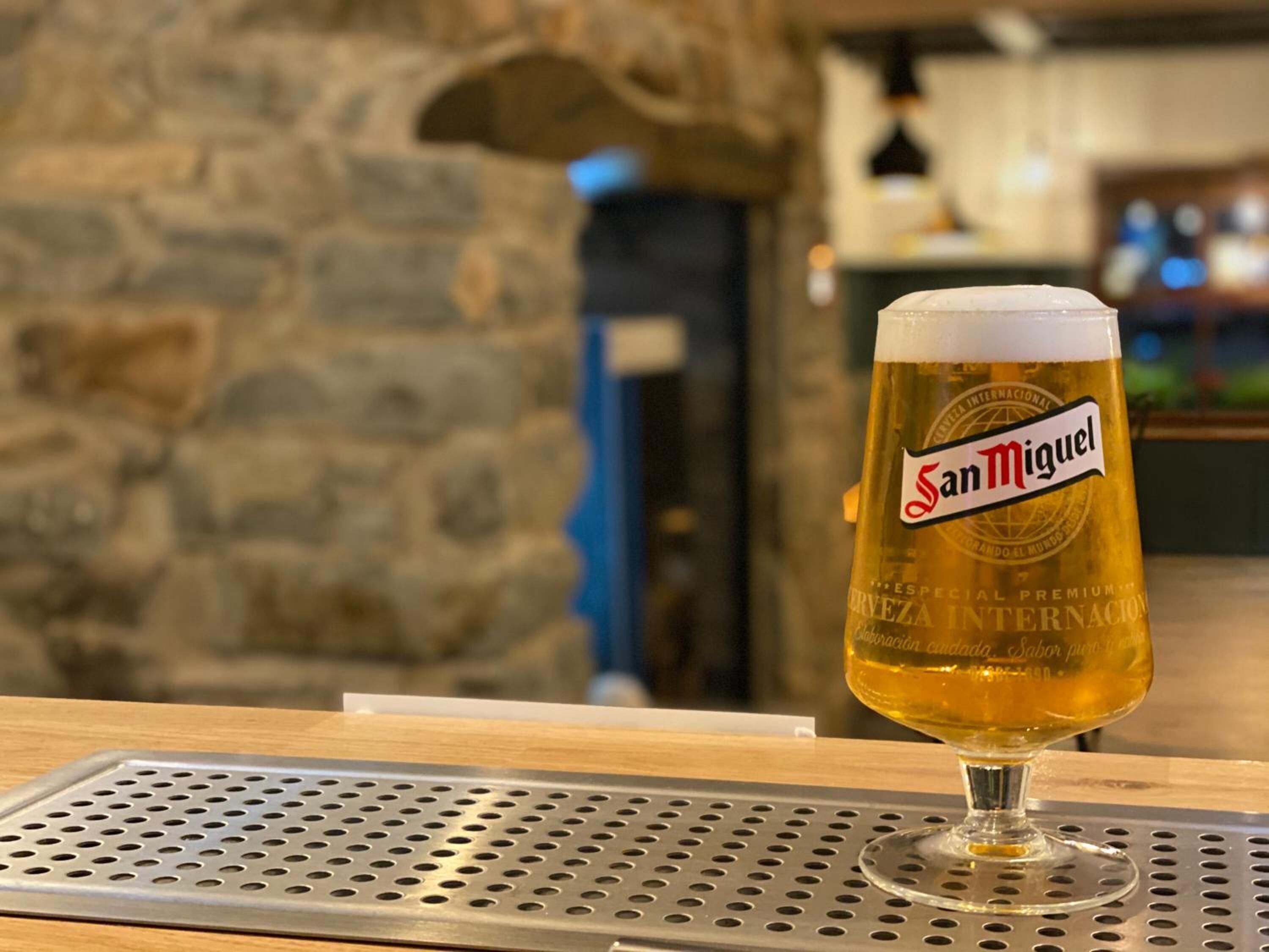 Un verre de bière San Miguel sur le comptoir d’un bar, avec un mur en pierre et une porte en arrière-plan.