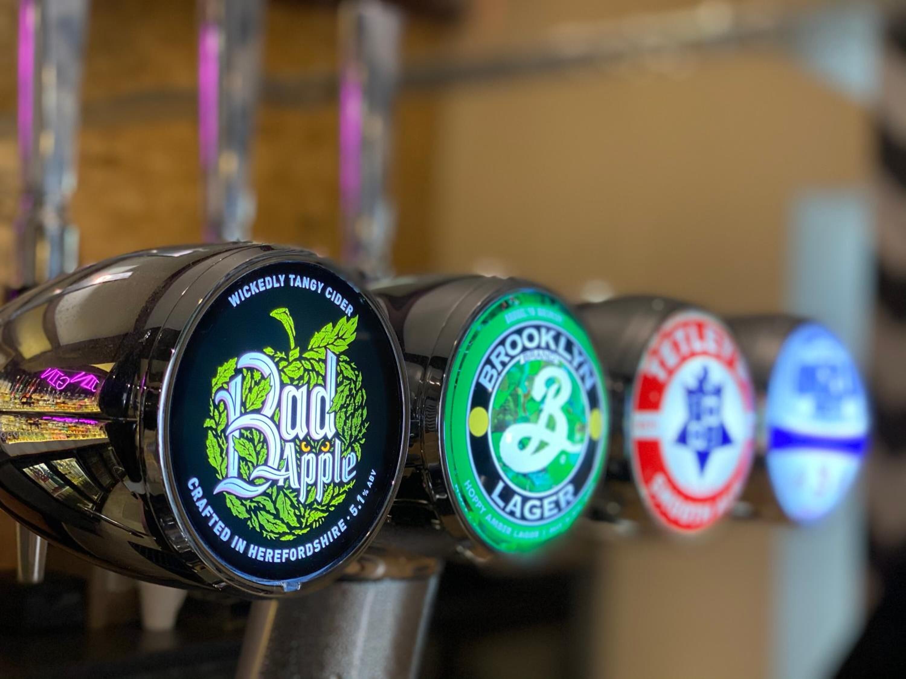 Un grifo con tres etiquetas de cerveza: Wickedly Tangy Cider, Brooklyn Lager y otra cerveza con etiqueta roja.