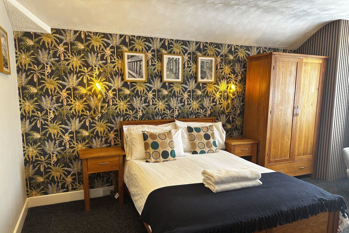 Un dormitorio con cama, mesitas de noche de madera, un armario y un papel tapiz tropical.