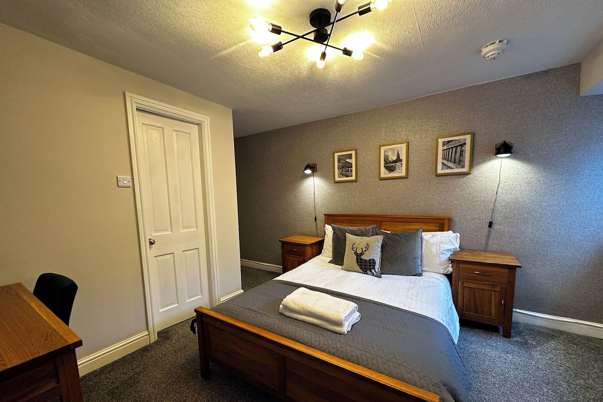 Un dormitorio con una cama de madera, un escritorio y una silla. Hay luces fijadas en la pared y cuadros enmarcados.