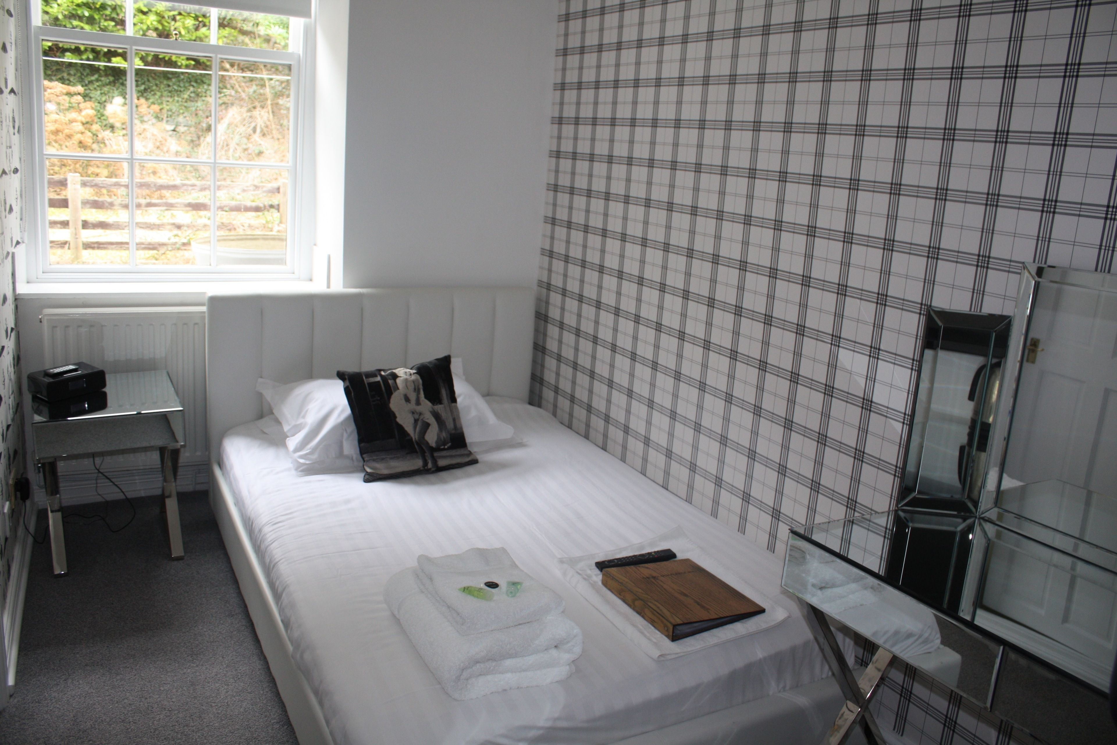 A neatly made bed with white linens, a bedside table with a lamp, and a window with a view of greenery outside.