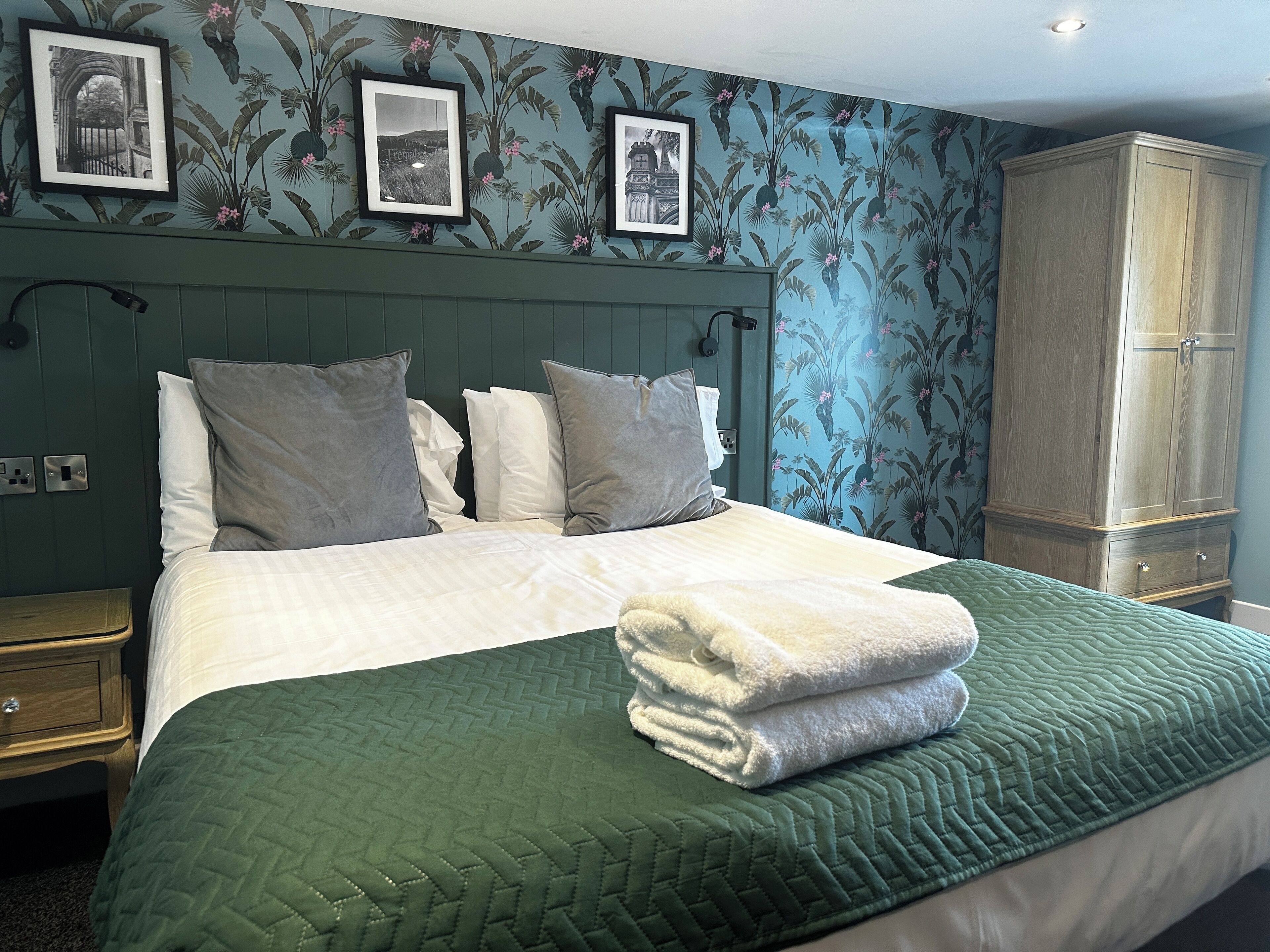 A neatly made bed with a green quilt, white pillows, and a folded white towel. There are framed pictures on the wall above the bed.