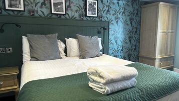 Una cama bien hecha con colcha verde, almohadas blancas y una toalla blanca doblada. Hay cuadros enmarcados en la pared sobre la cama.