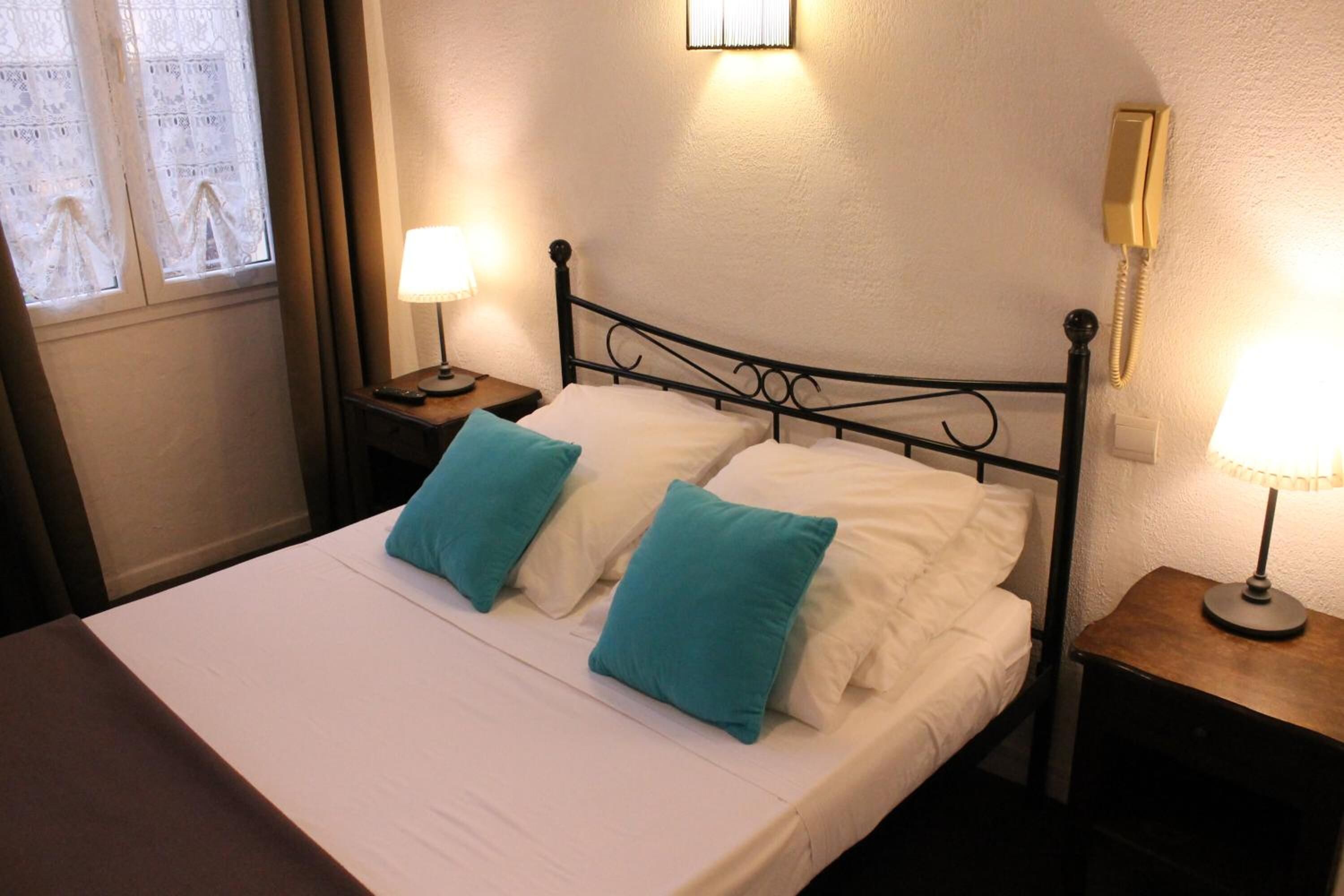 A neatly made bed with white and teal pillows, a bedside table with a lamp, and a phone on the wall.