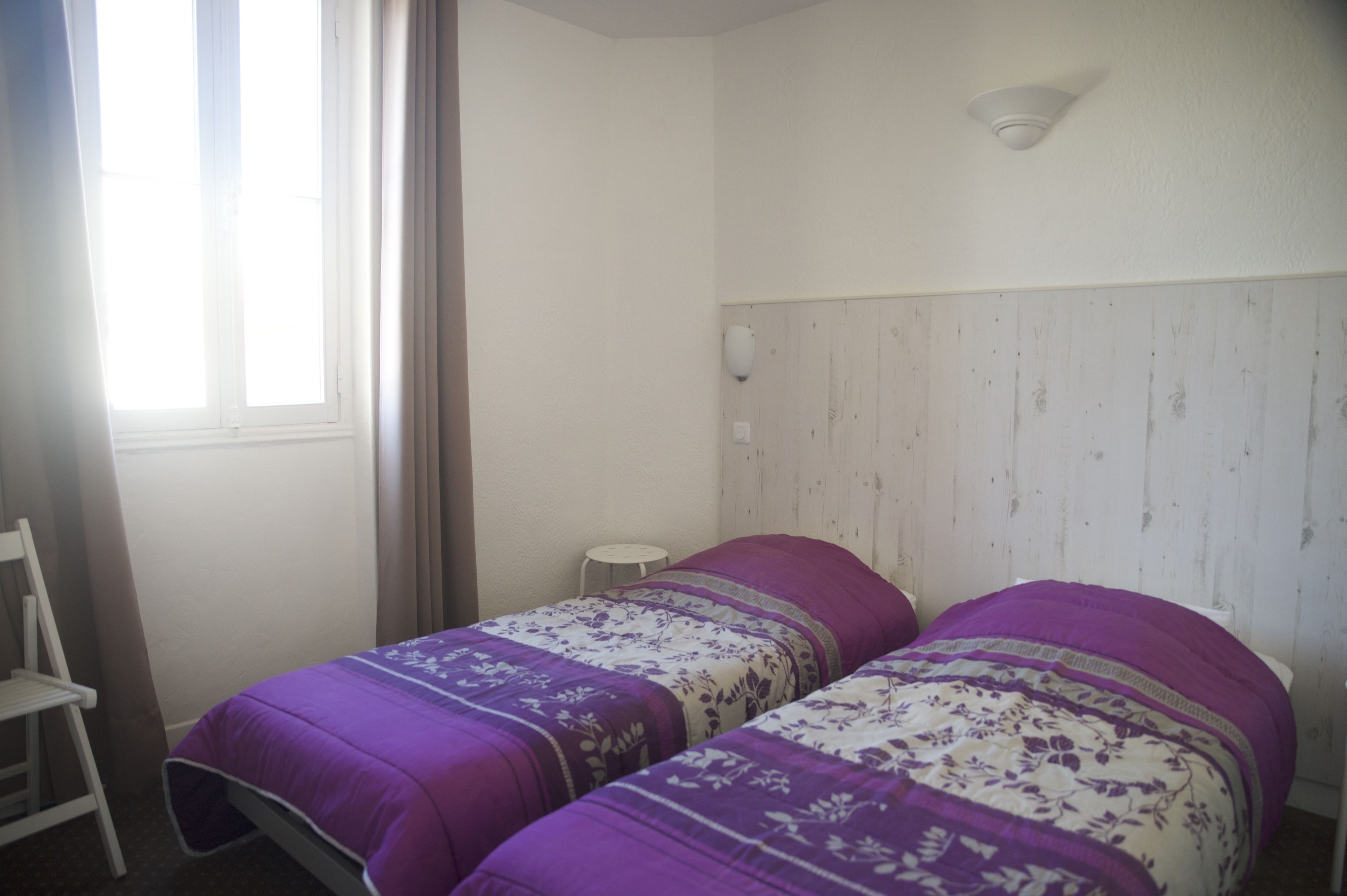 Een eenvoudige kamer met twee bedden, een stoel, een klein tafeltje en een raam met gordijnen.