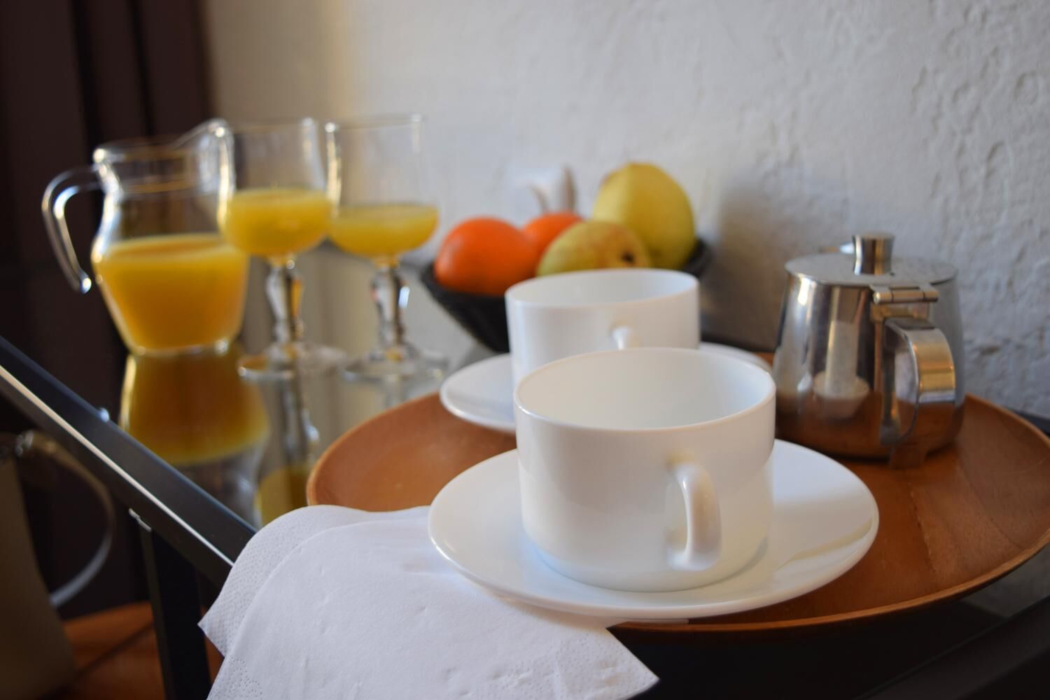 A breakfast tray with white cups, a pitcher of orange juice, and a fruit bowl containing apples and oranges.
