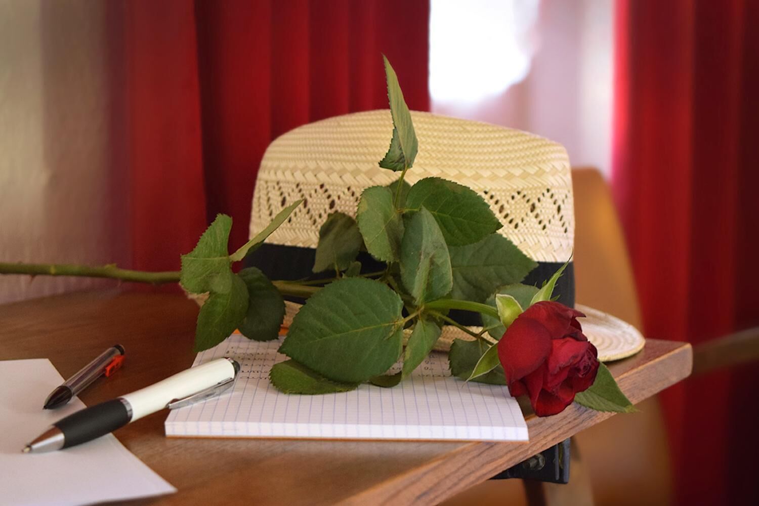 Un cuaderno, un bolígrafo, una rosa y un sombrero tejido sobre una mesa de madera.
