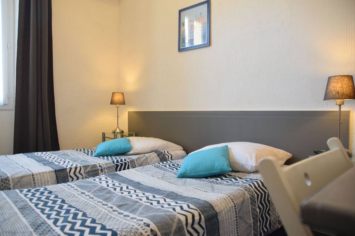 A hotel room with two beds, each with patterned bedding and pillows, a nightstand with a lamp, a chair, and a framed picture on the wall.