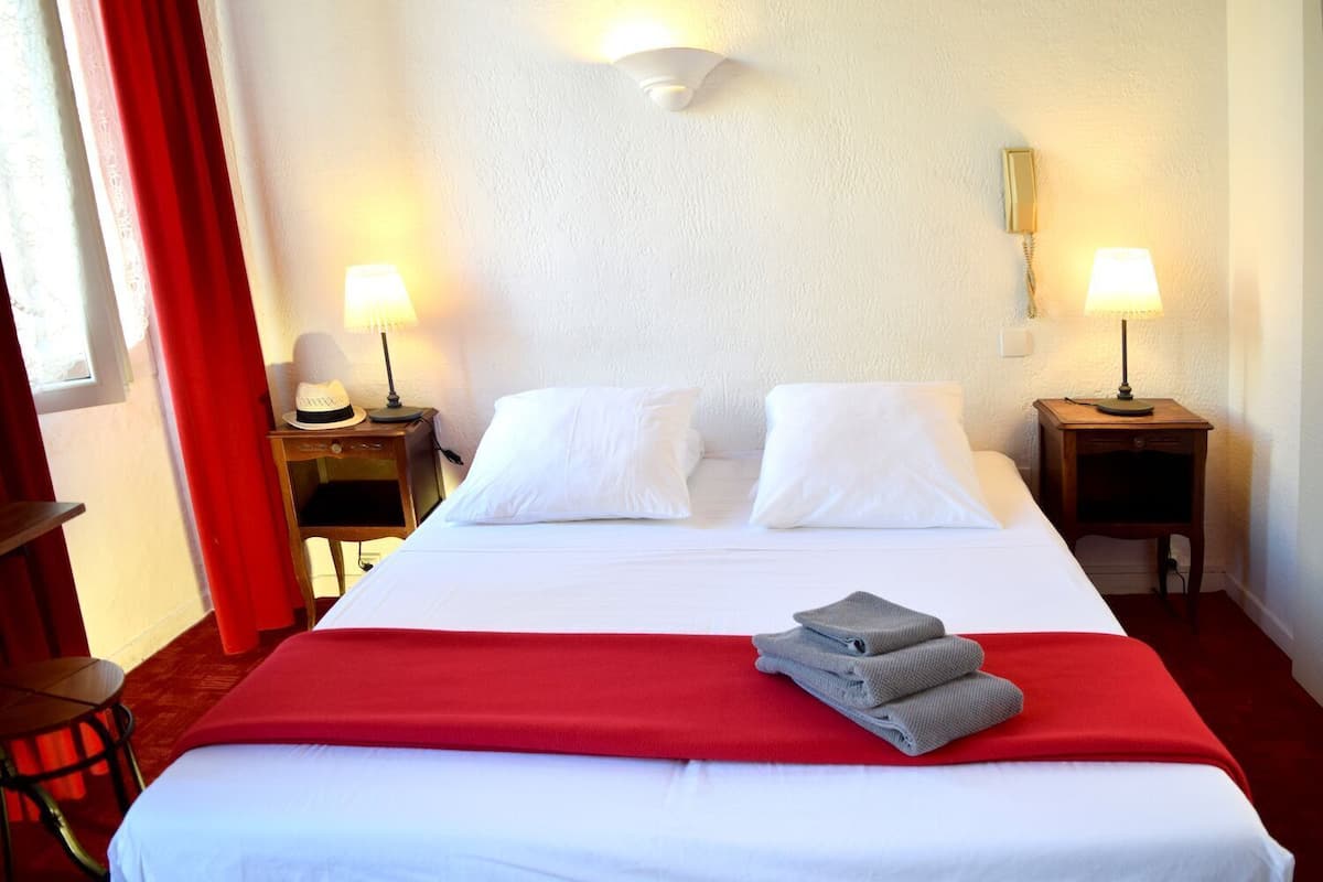 A hotel room with a bed, two bedside lamps, a red curtain, and a red throw blanket.