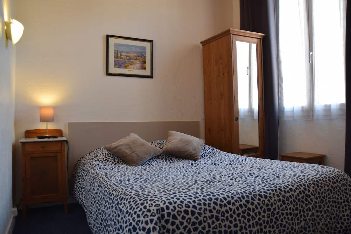 A bedroom with a bed, a nightstand, a wooden wardrobe, a framed picture on the wall, and a window with curtains.