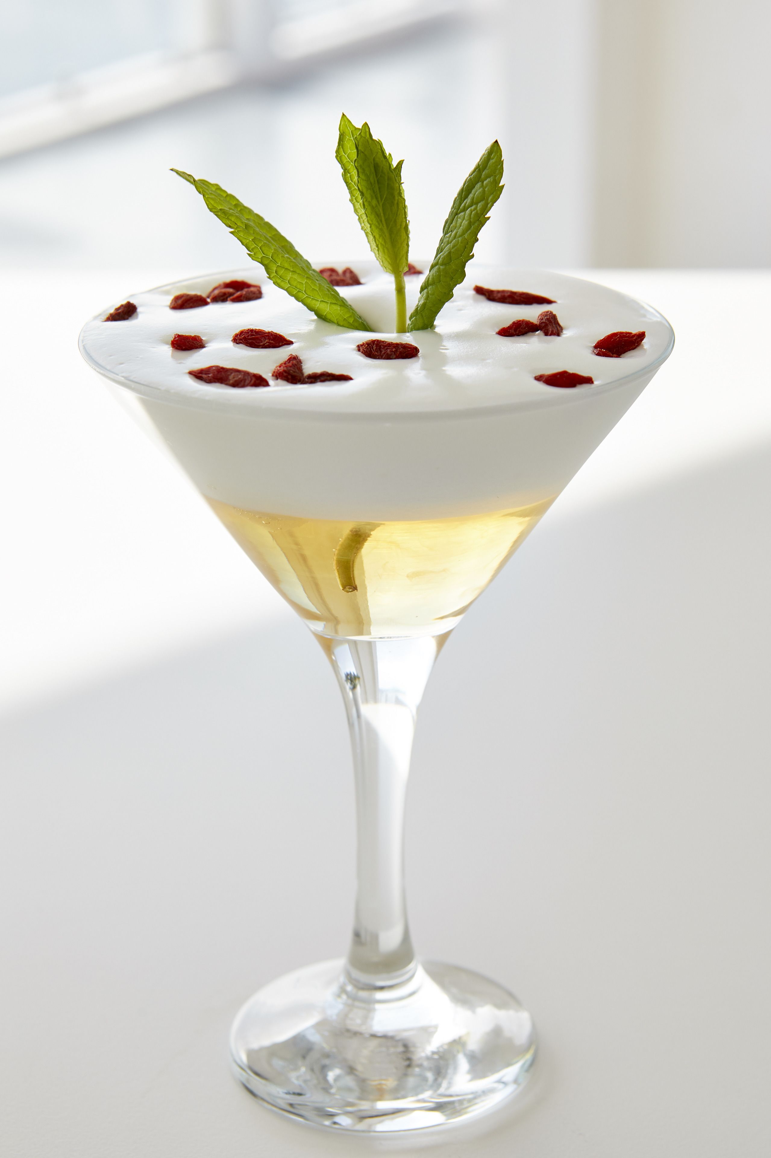 Una copa de martini con una capa blanca y cremosa, adornada con hojas de menta y bayas rojas, sobre una superficie blanca.