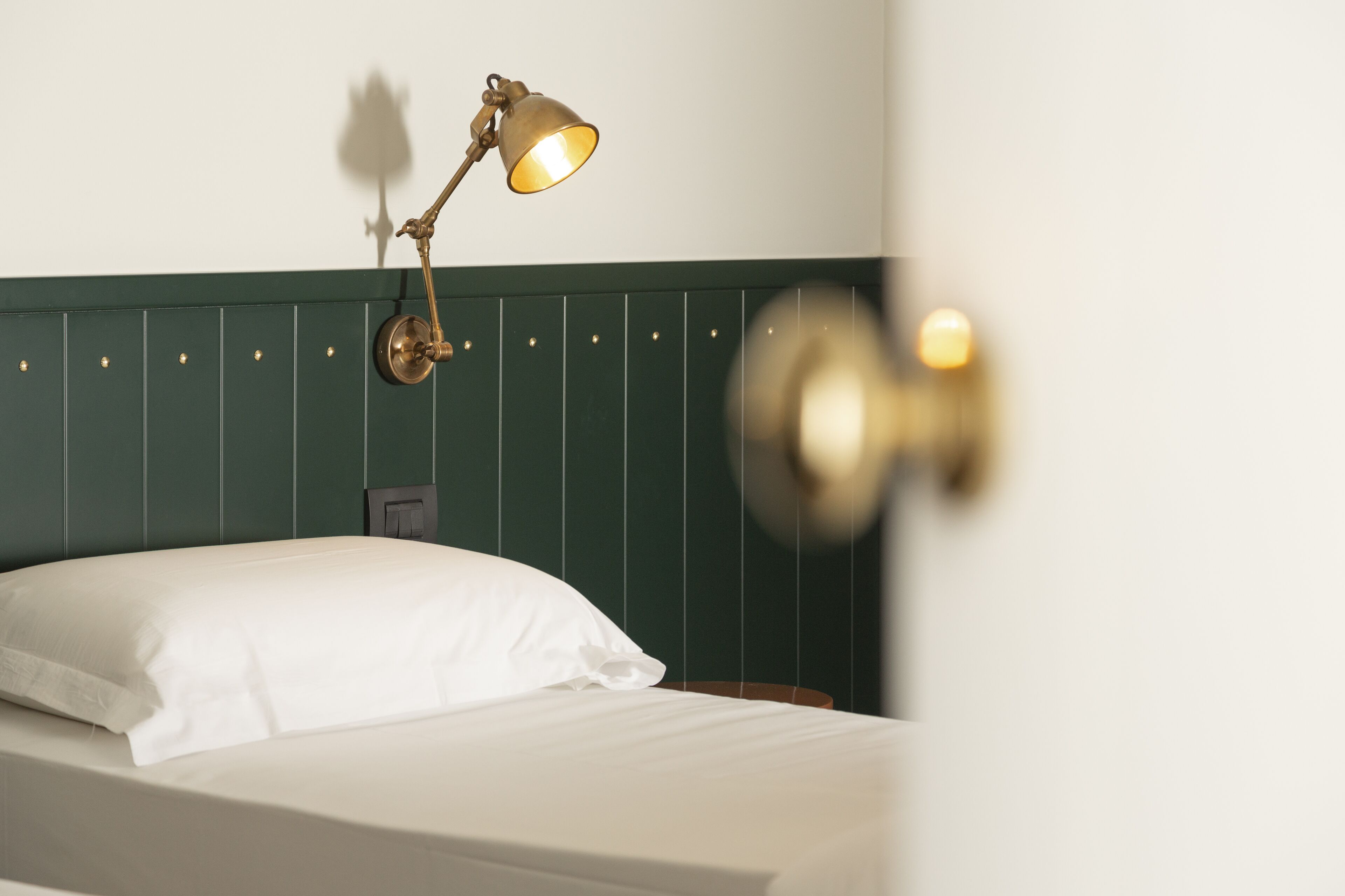 A neatly made bed with white linens and a single white pillow. A brass wall sconce with a warm light illuminates the room.