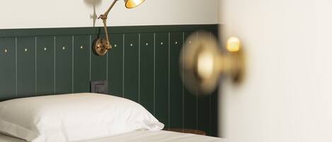 A neatly made bed with white linens and a single white pillow. A brass wall sconce with a warm light illuminates the room.