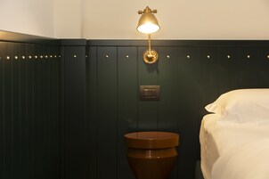 Una habitación de hotel con una cama, una mesita de noche, una lámpara de pared y un panel con un interruptor.