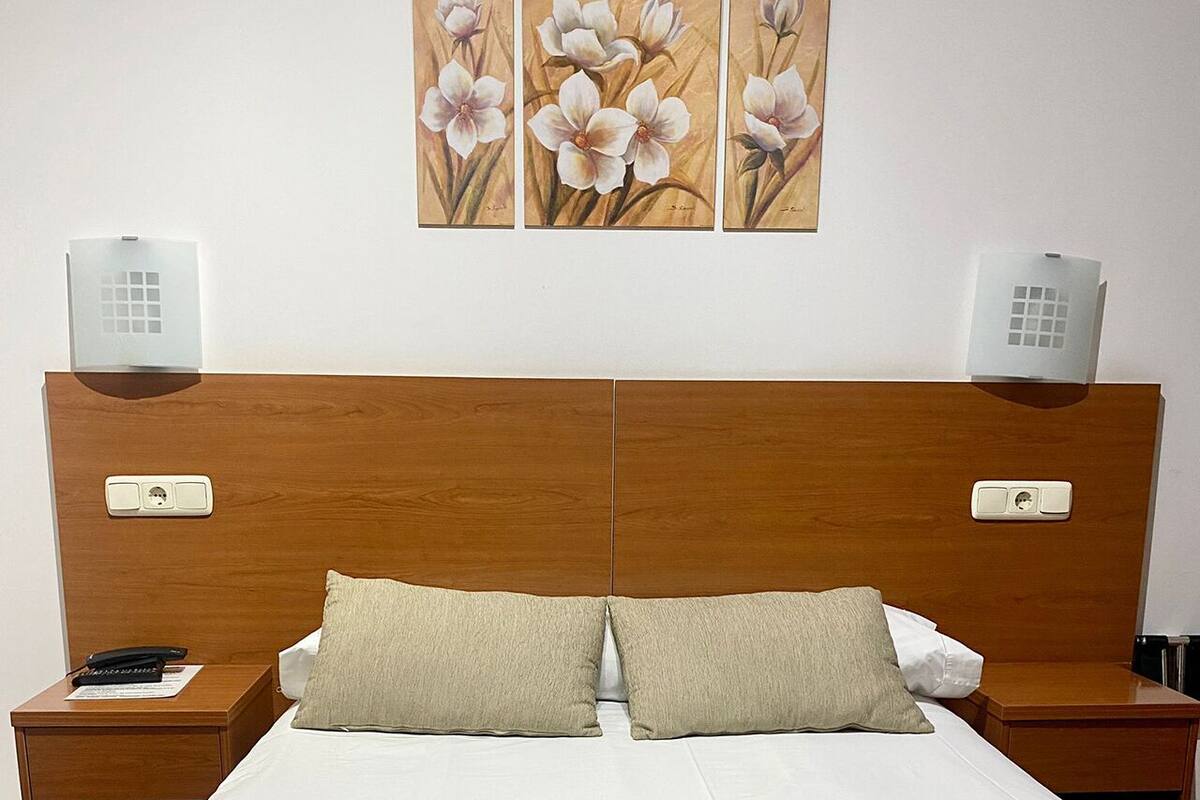 Habitación de hotel con cabecera de madera, dos mesitas de noche, cama con ropa blanca, dos almohadas color beige y una manta de color beige. Hay tres apliques de pared y un cuadro de flores sobre la cama.