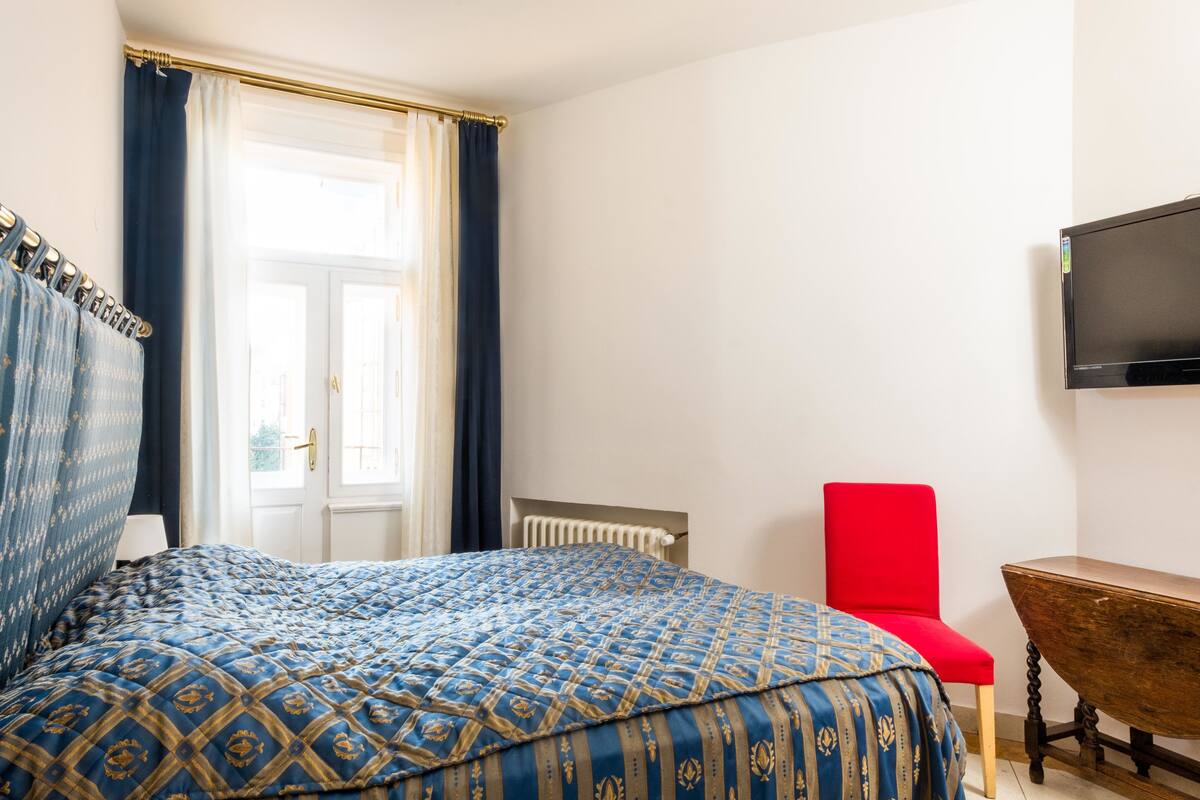 A bedroom with a bed, a red chair, a wooden table, and a TV on the wall.