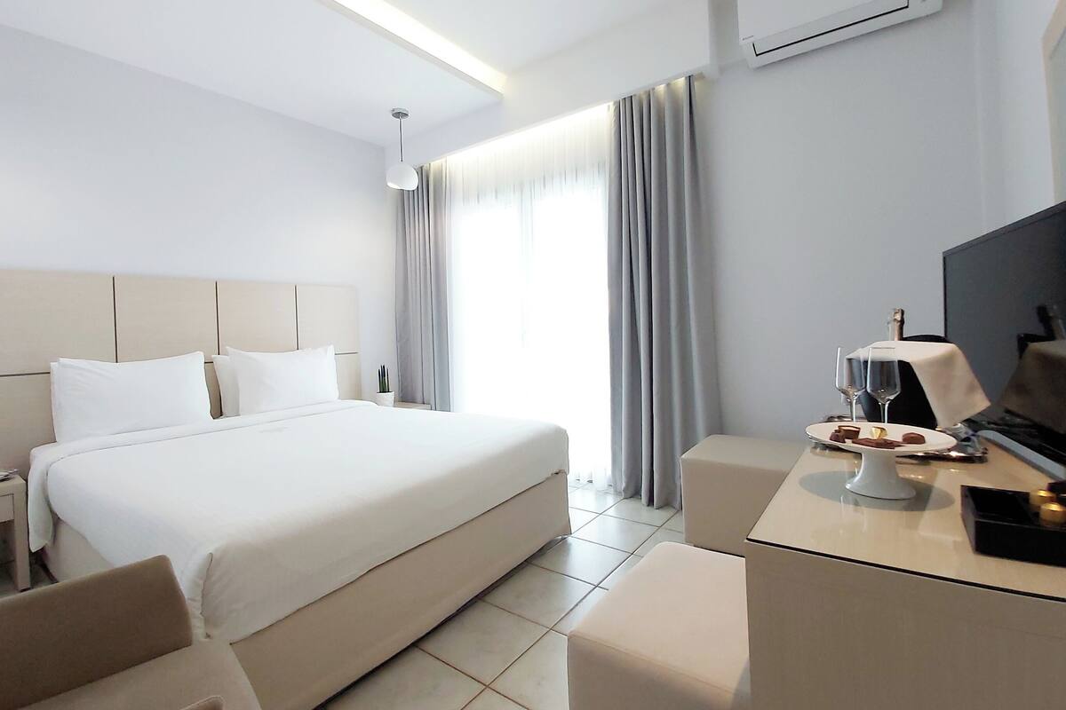 A modern hotel room with a large bed, a desk with a television, and a small table with a bottle and glasses.